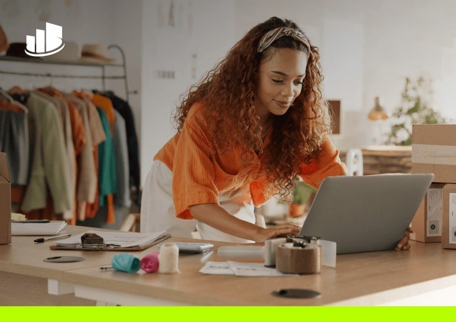 A young woman with curly hair works on a laptop in a boutique-style workspace surrounded by clothing racks, shipping boxes, and packaging materials, suggesting a small retail or e-commerce business.