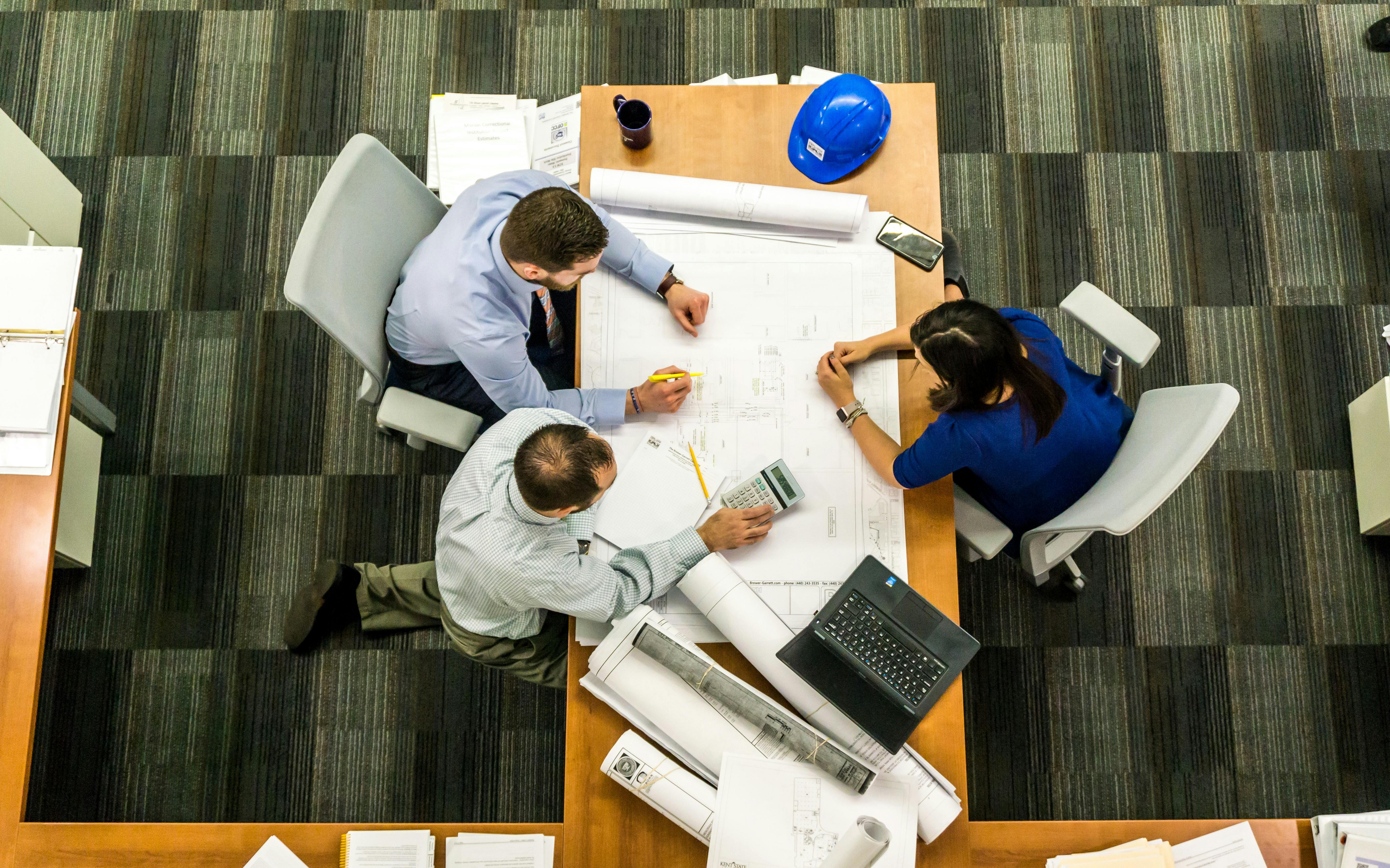 Business Continuity. Three individuals sitting at a table, examining blueprints and discussing plans.
