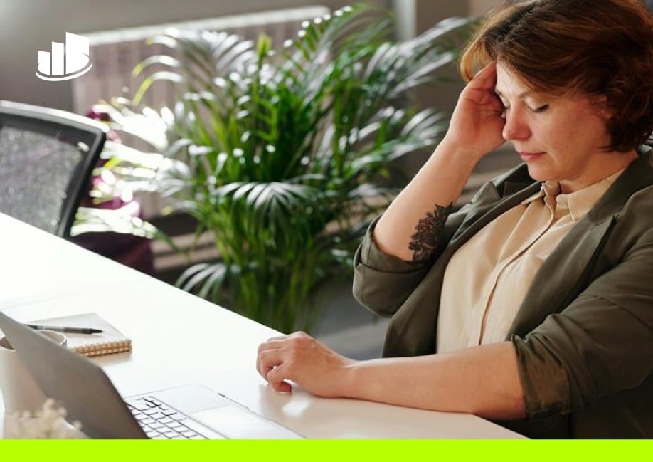 A woman in a blazer sits at a desk with a laptop appearing stressed as she faces a challenge. 