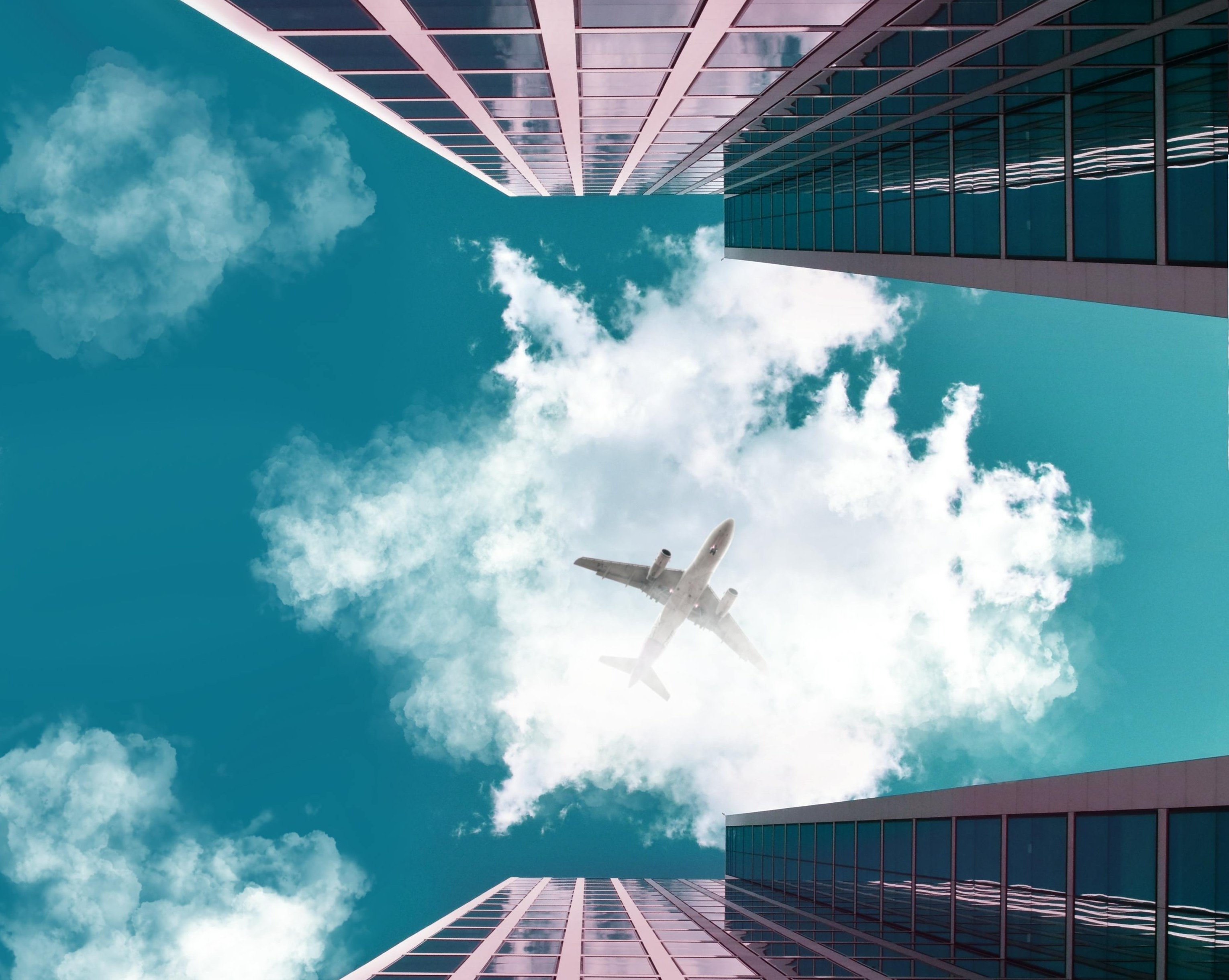 Looking up from between two skyscrapers at a passing airplane against a backdrop of blue sky and fluffy clouds.